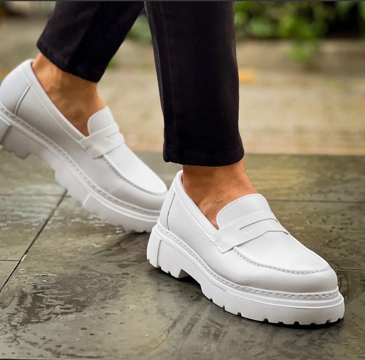 White leather chunky loafers worn with black trousers on a stone pavement.