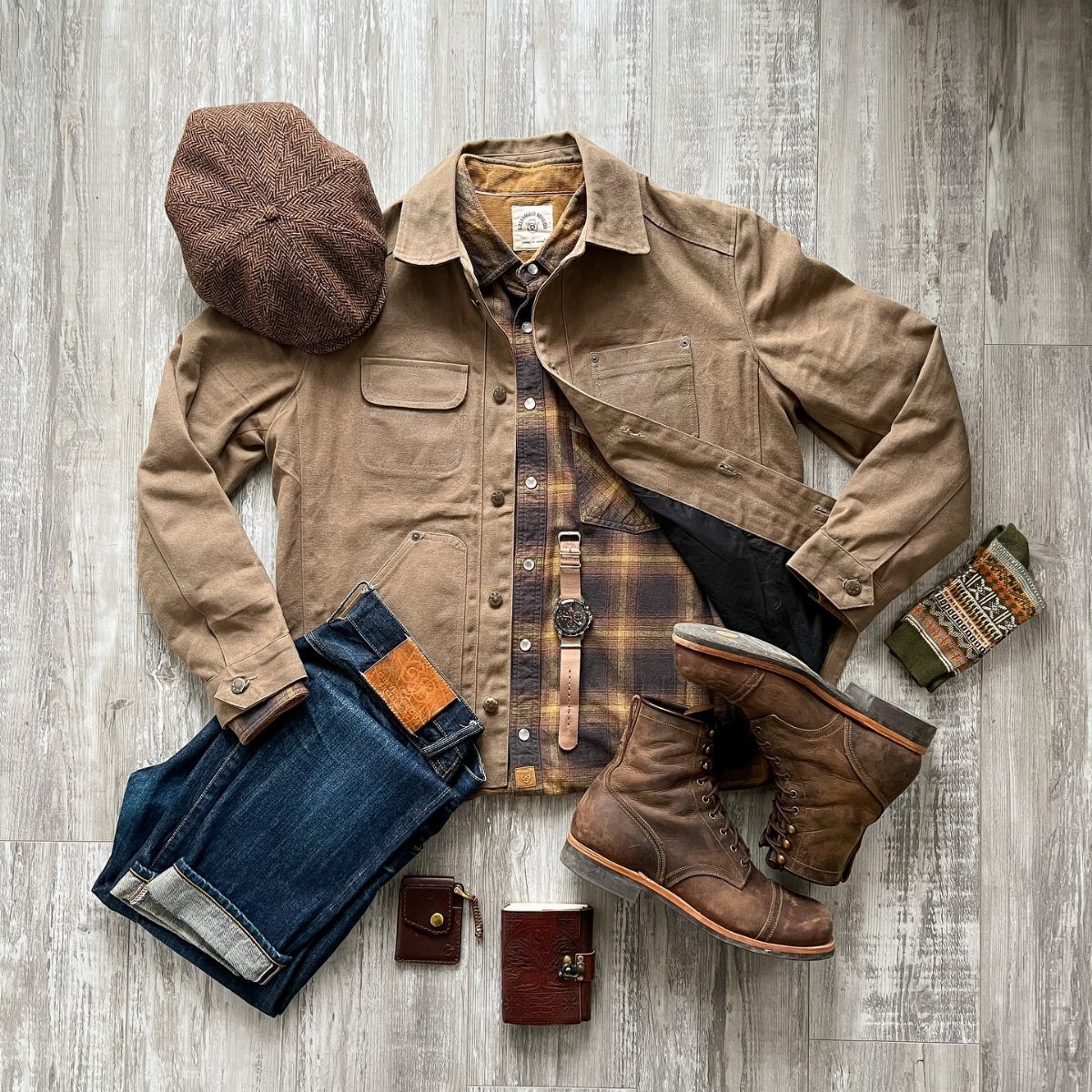 Men's rugged clothing flat lay featuring a tan jacket, plaid shirt, and leather boots.
