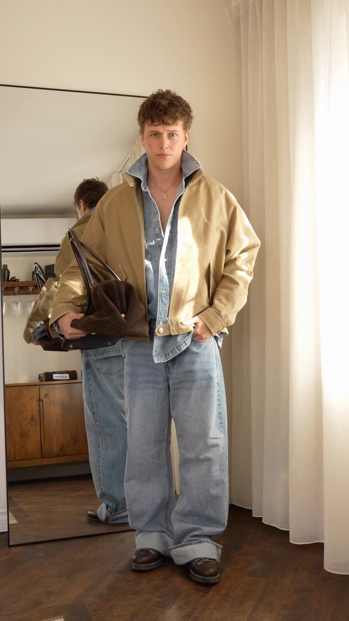 Man wearing a tan jacket, blue shirt, and wide denim jeans holding a bag.