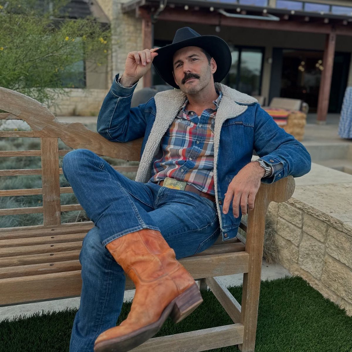 Man in cowboy hat, denim jacket, and leather boots sitting on wooden bench.
