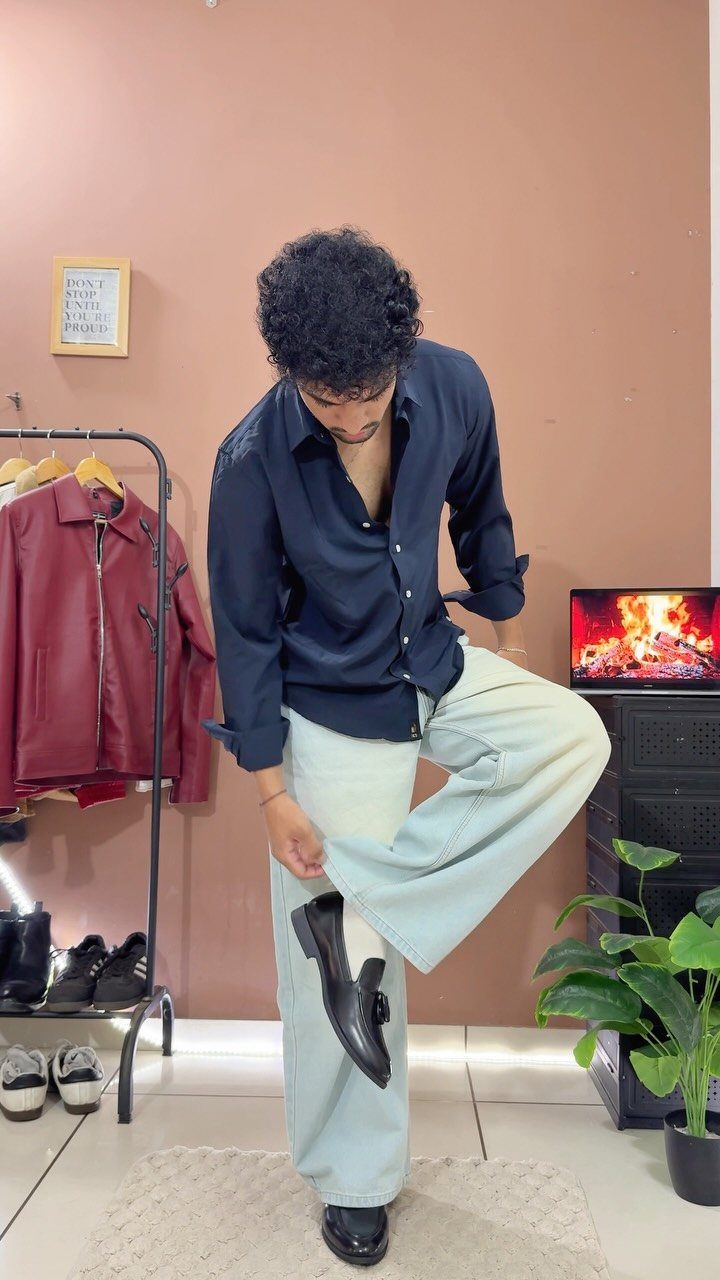Man wearing light-colored wide-leg jeans putting on black leather dress shoes indoors.