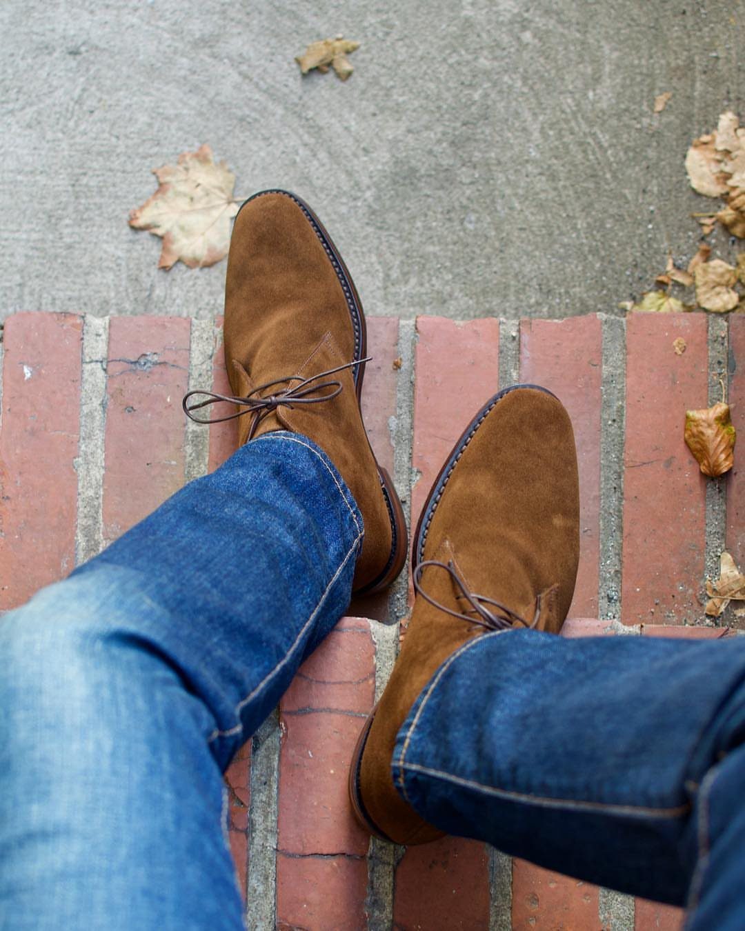 Brown suede chukka boots paired with blue denim jeans on red brick pavement.