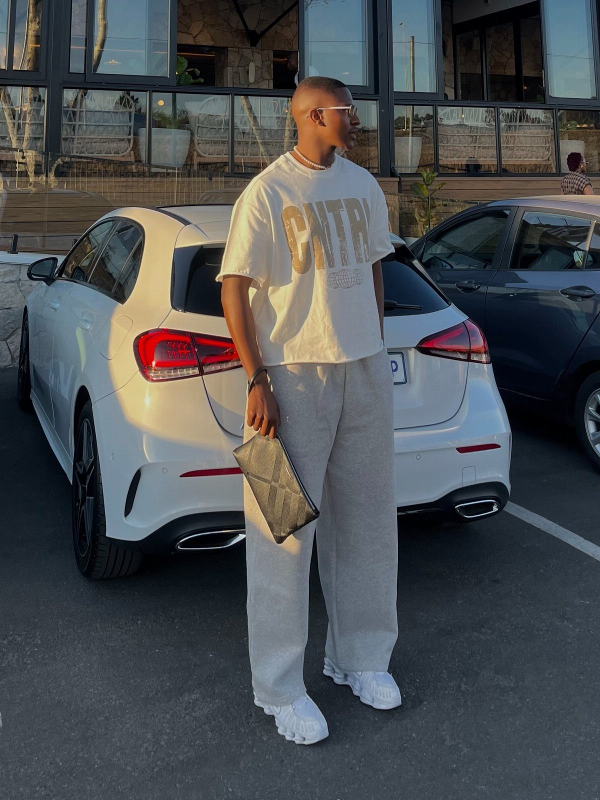 Man in cream t-shirt and grey sweatpants wearing white sneakers by a car