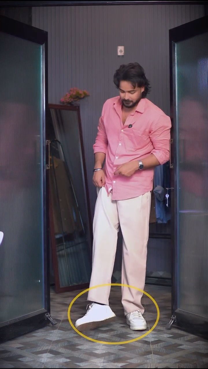 Man in pink shirt and cream trousers wearing white sneakers in a room.