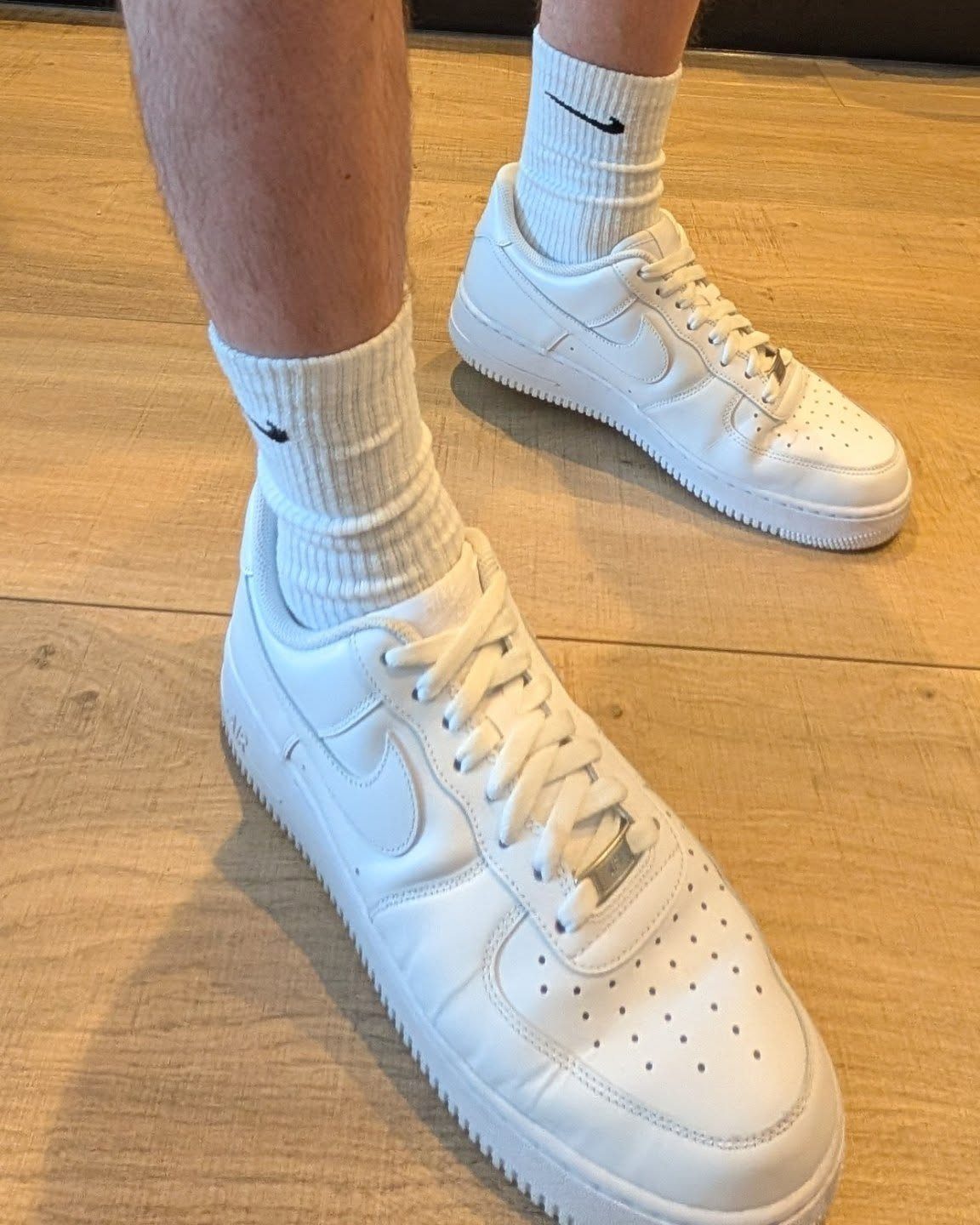 White Nike sneakers and white crew socks on a light wooden floor.
