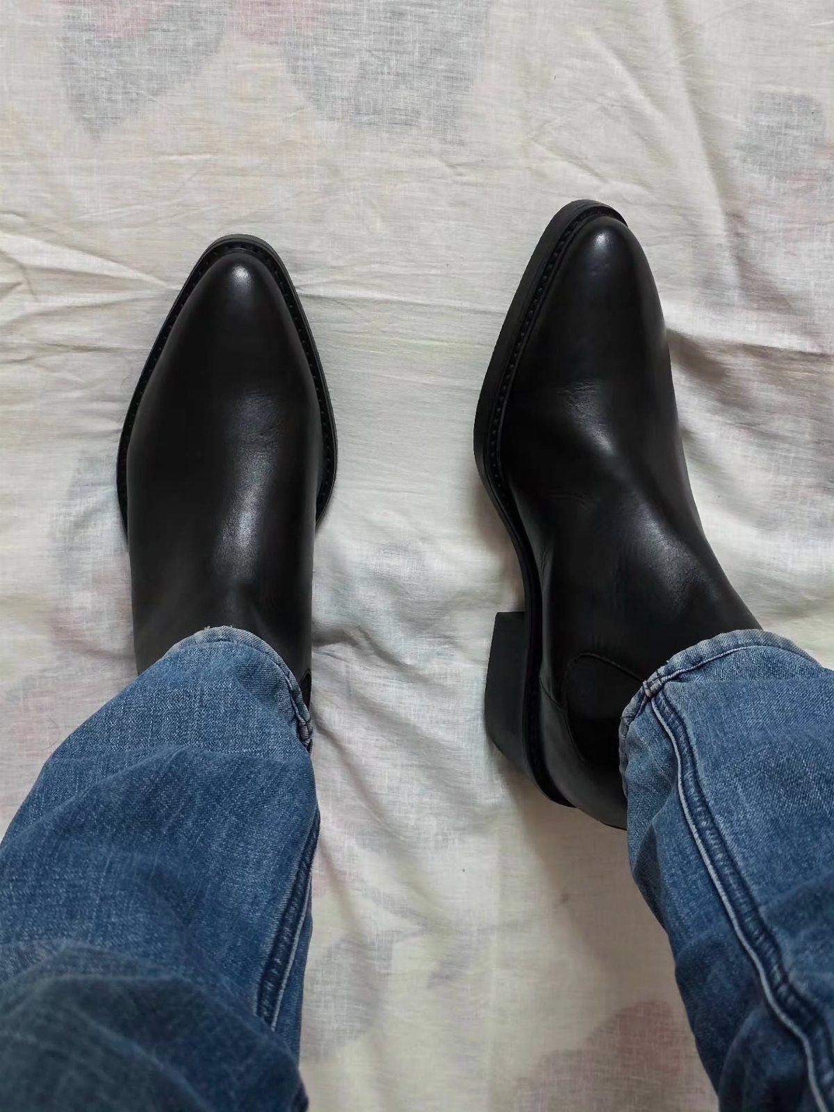 Black leather boots worn with blue jeans on a light fabric background.