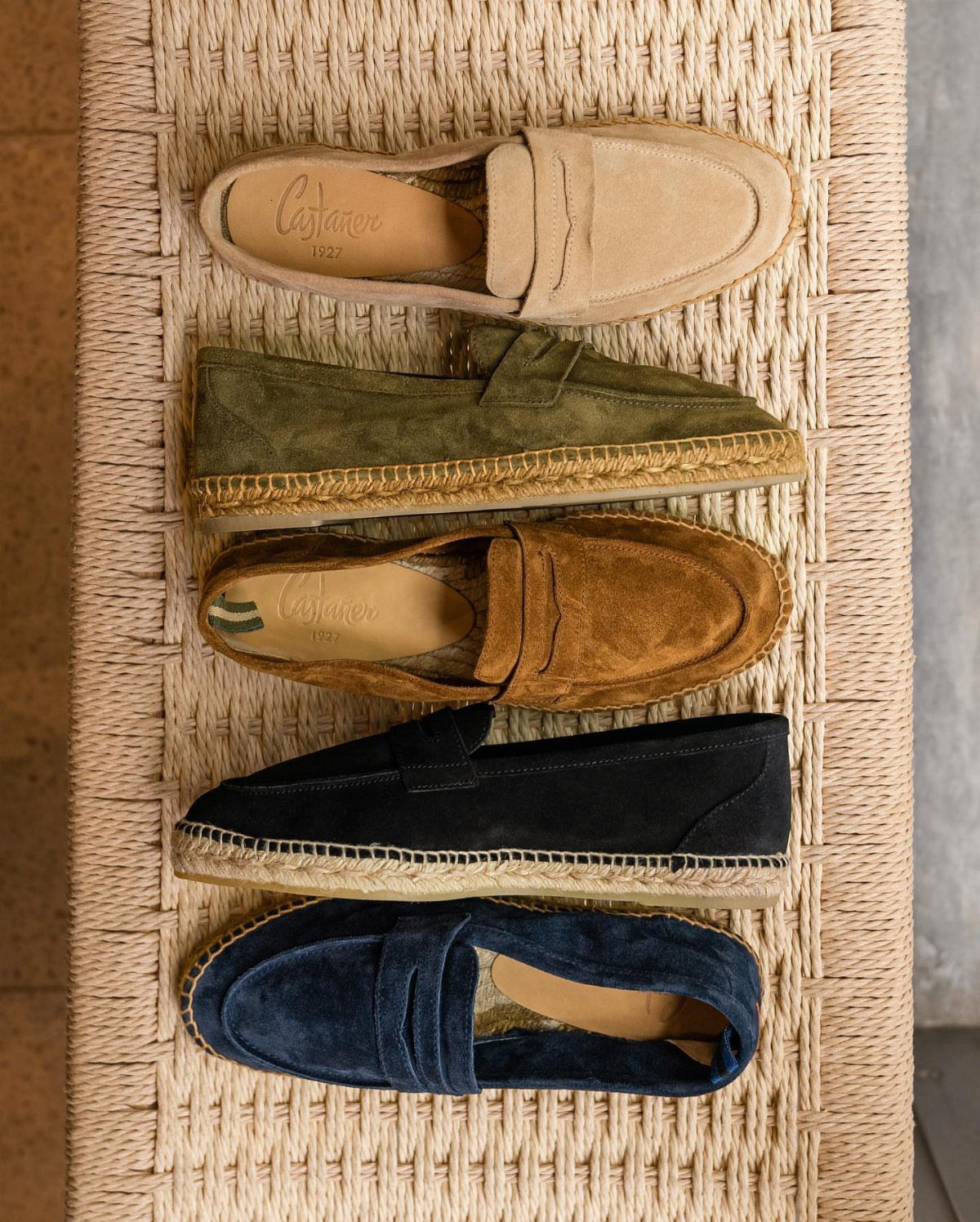 Five pairs of colorful suede espadrilles arranged on a woven straw mat.