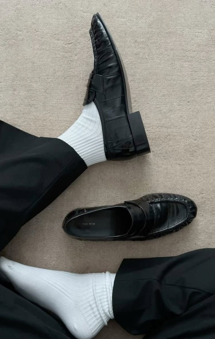 Black leather loafers with white socks and black trousers on grey carpet.