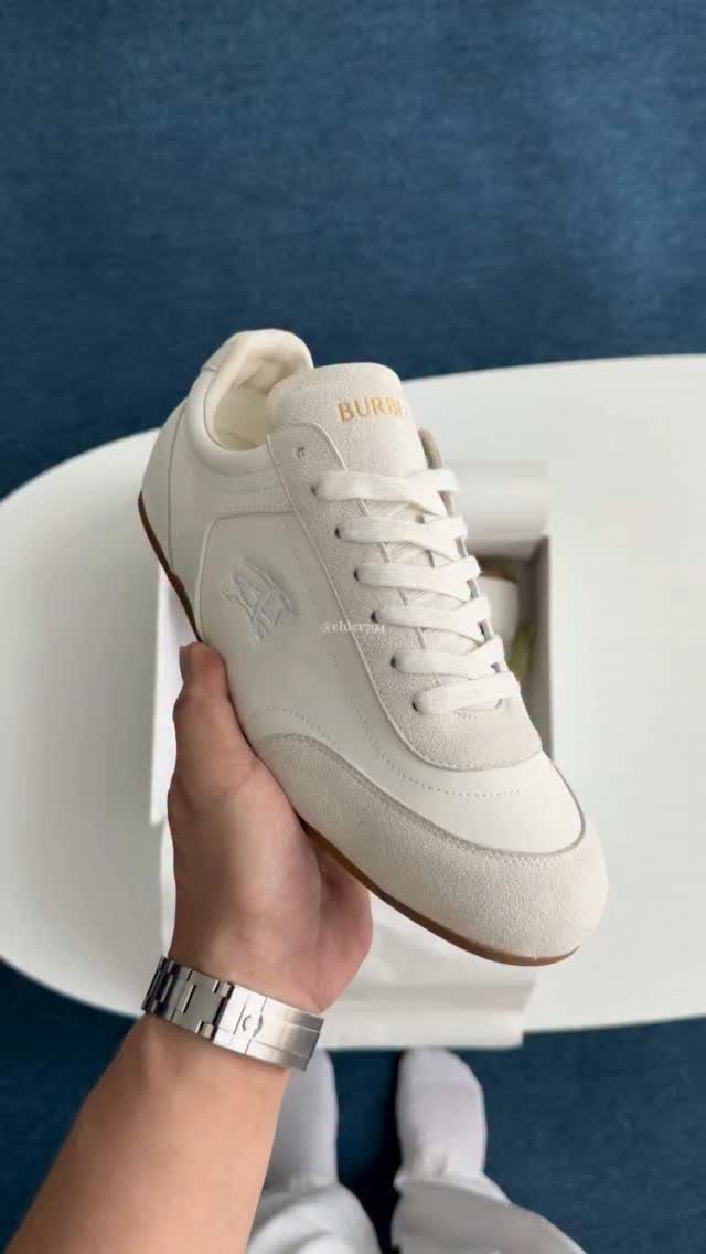 Cream-colored suede sneaker held by a hand over a white shoe box.
