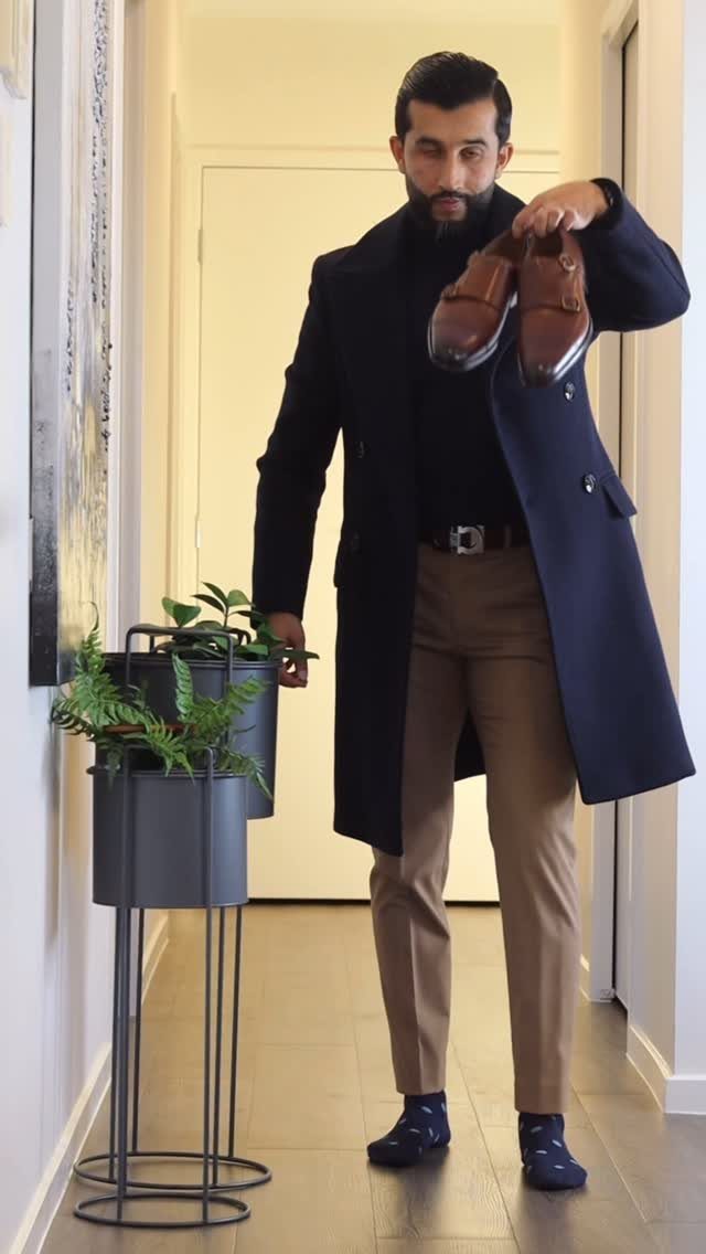 Man in navy coat holding brown leather monk strap dress shoes in hallway