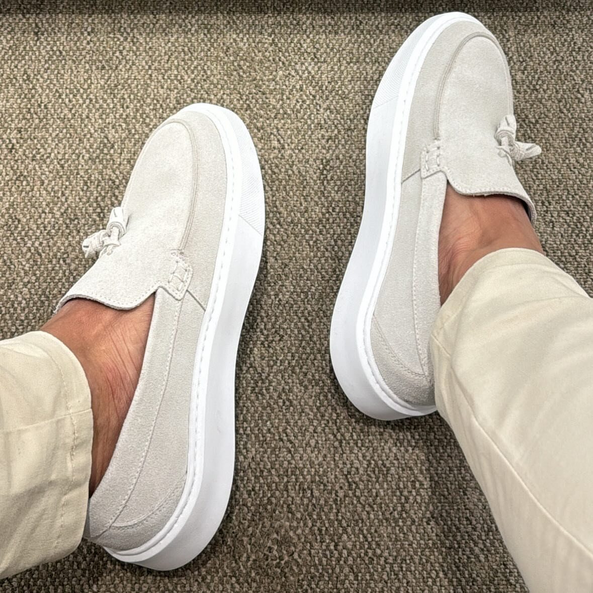 Beige suede slip-on shoes with white soles and tassels on a brown carpet.