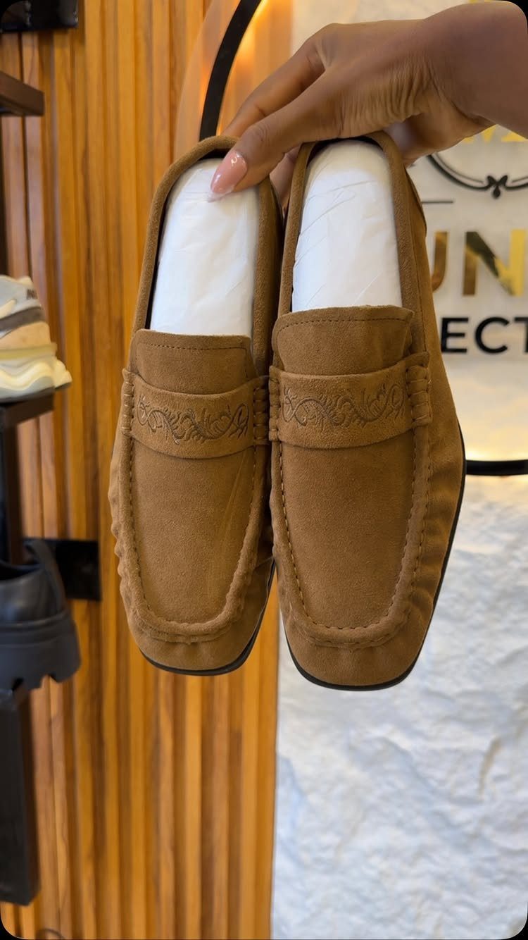 Tan suede moccasin shoes held up by a hand against a wooden background.