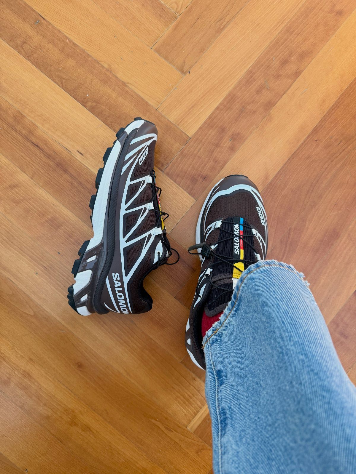 Brown and white Salomon sneakers worn with blue jeans on a wooden floor