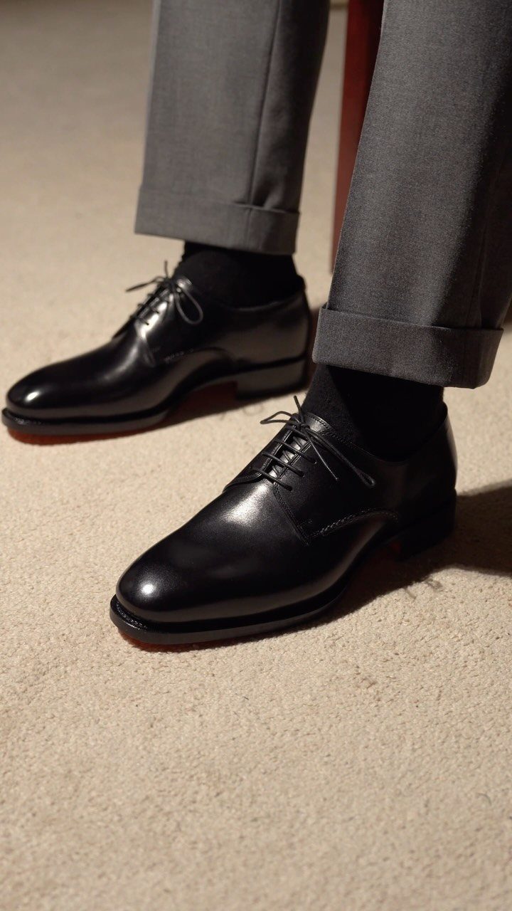 Black leather Oxford shoes worn with grey trousers and black socks on carpet.
