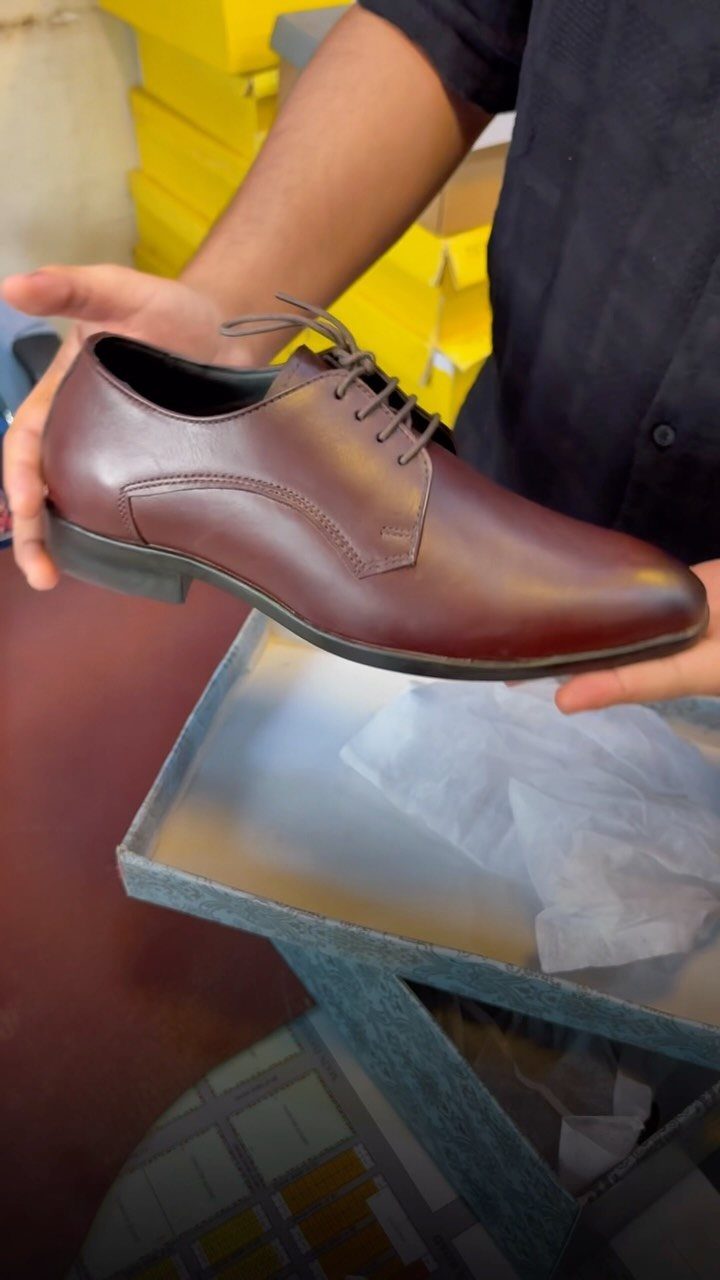 Brown leather derby shoe held by hands against a background of yellow boxes.