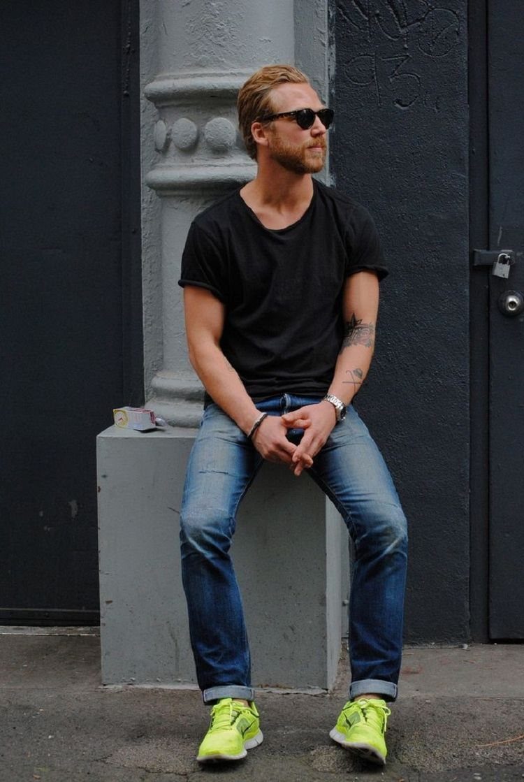 Man wearing black t-shirt, blue jeans, and bright yellow sneakers sitting outdoors.