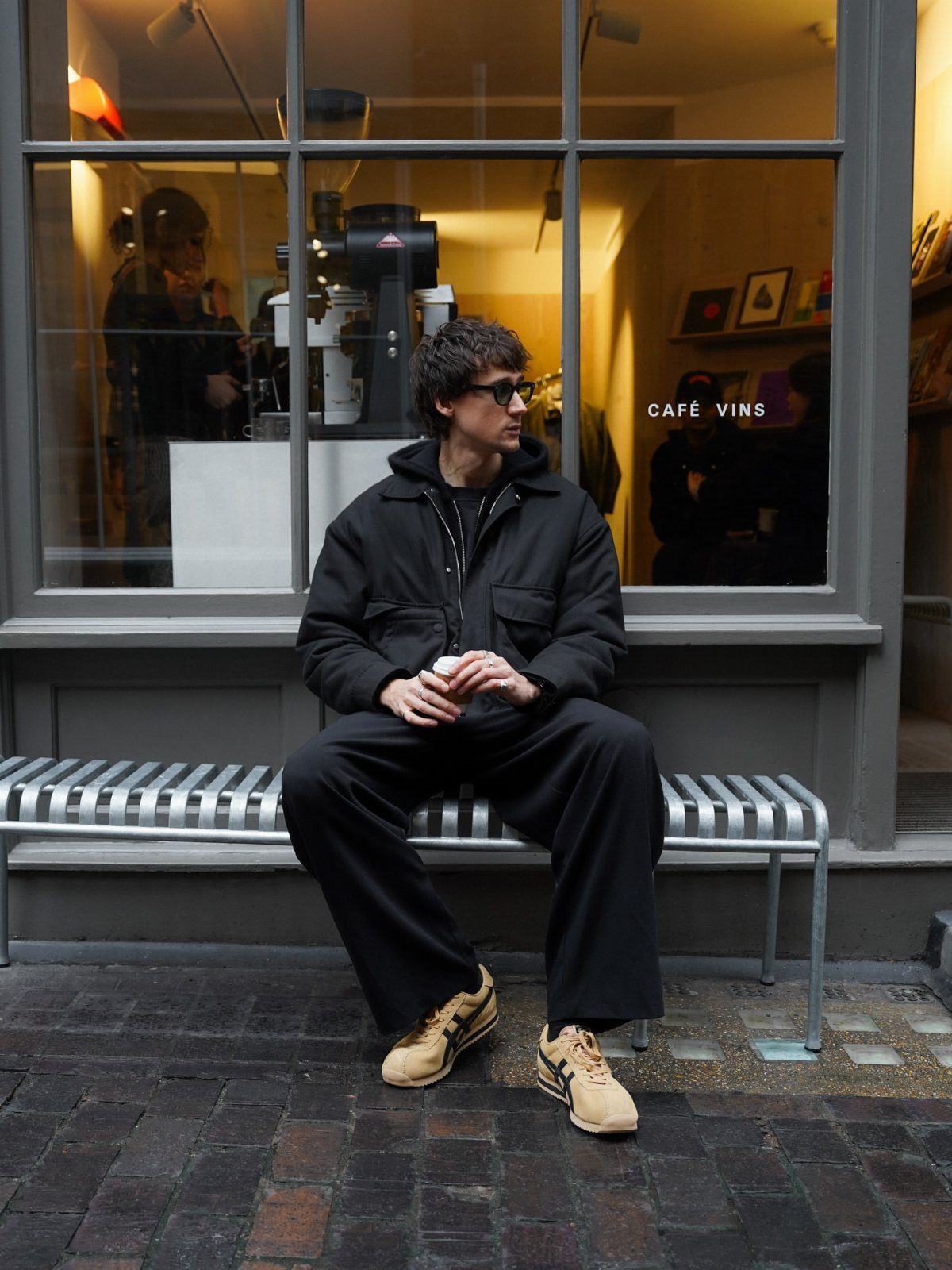Man in black outfit and yellow sneakers sitting on a metal bench outdoors.