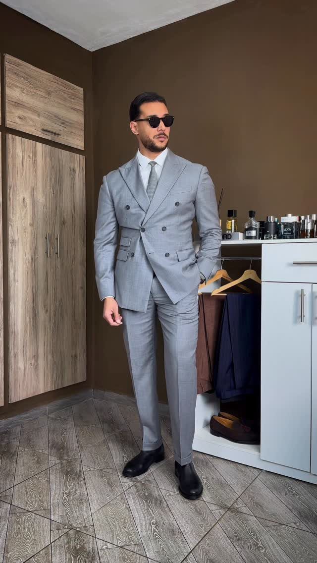 Man in a grey double-breasted suit and black Chelsea boots standing indoors.