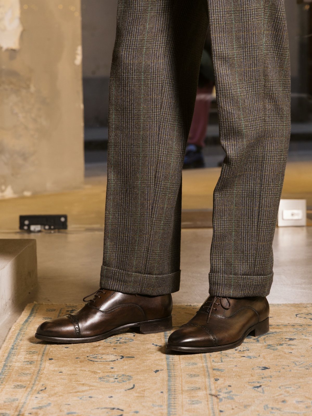 Brown leather oxford shoes paired with cuffed grey textured wool trousers on a rug.