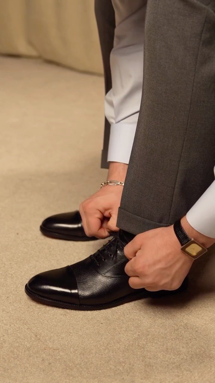 Person tying laces of black leather oxford shoes while wearing grey dress trousers.