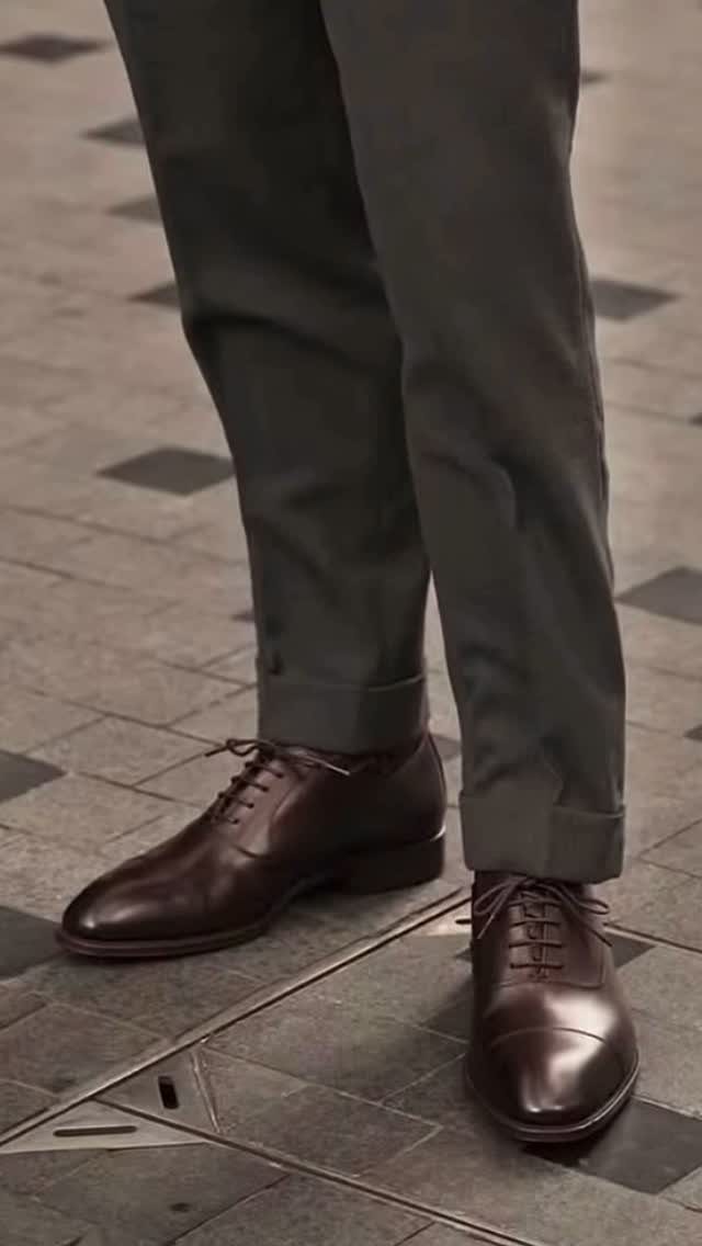 Brown leather oxford shoes paired with grey cuffed trousers on a tiled floor.