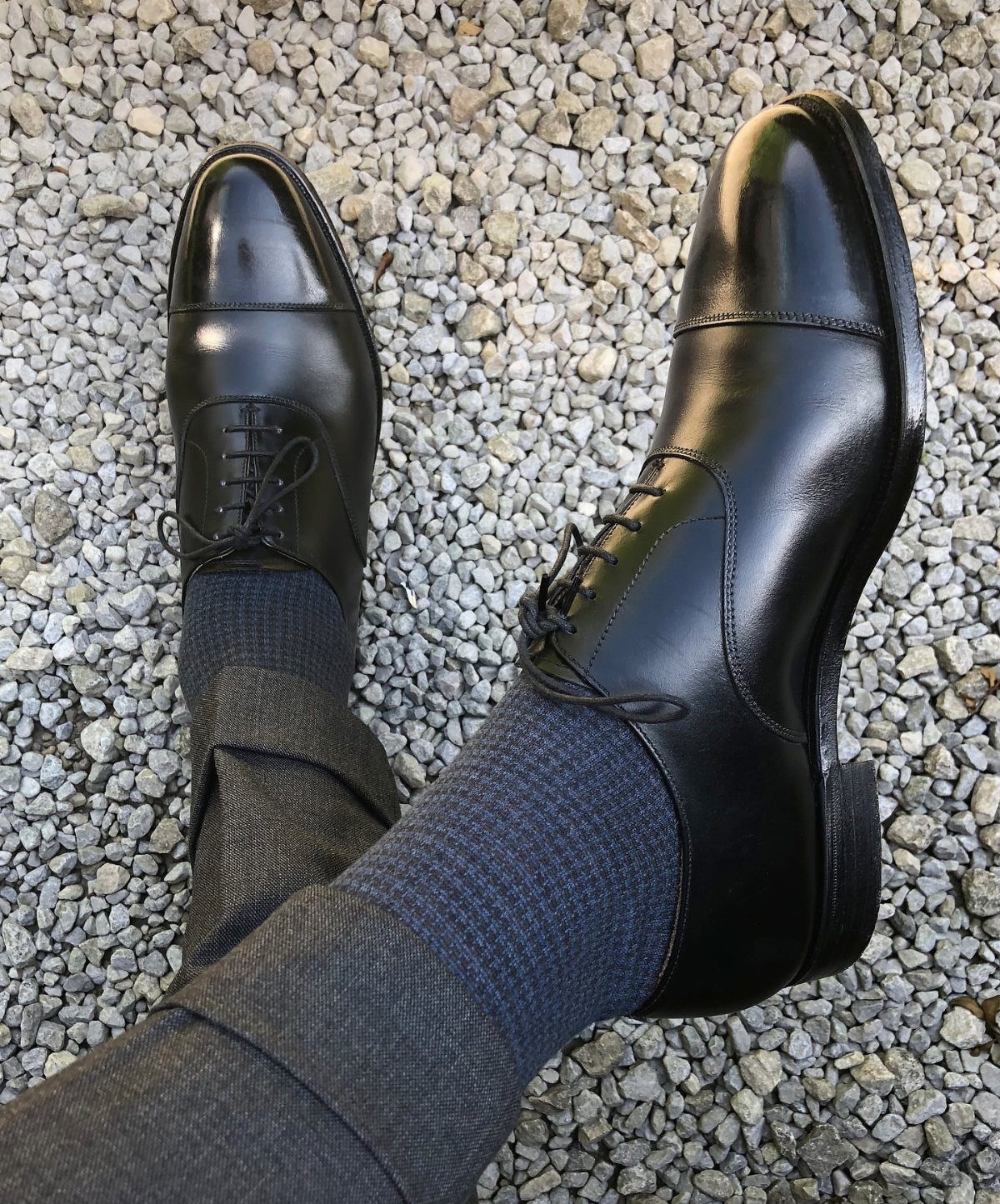Black leather whole cut oxford shoes worn with grey trousers and socks on gravel