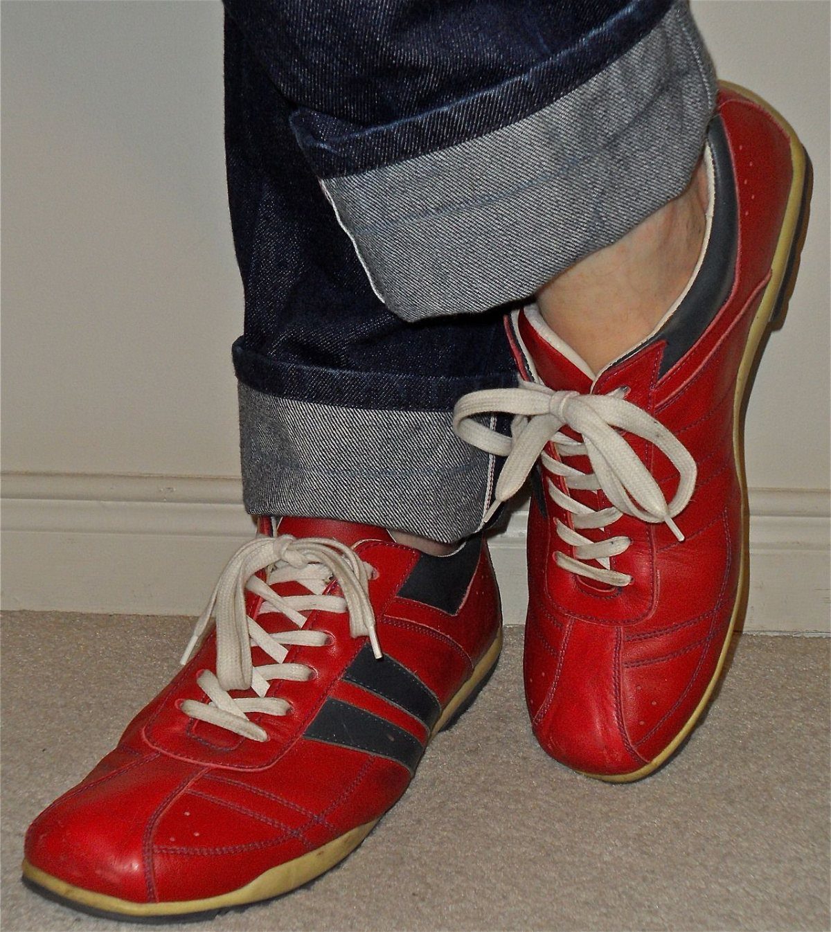 Red leather sneakers with white laces paired with dark blue cuffed denim jeans.