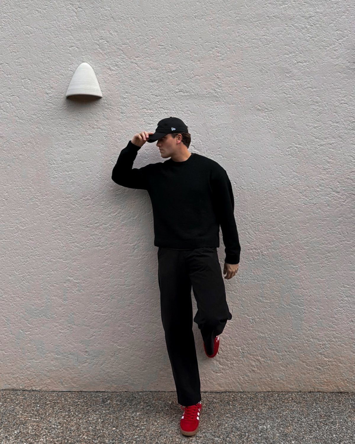 Man in all-black streetwear outfit wearing bright red sneakers against a grey wall.