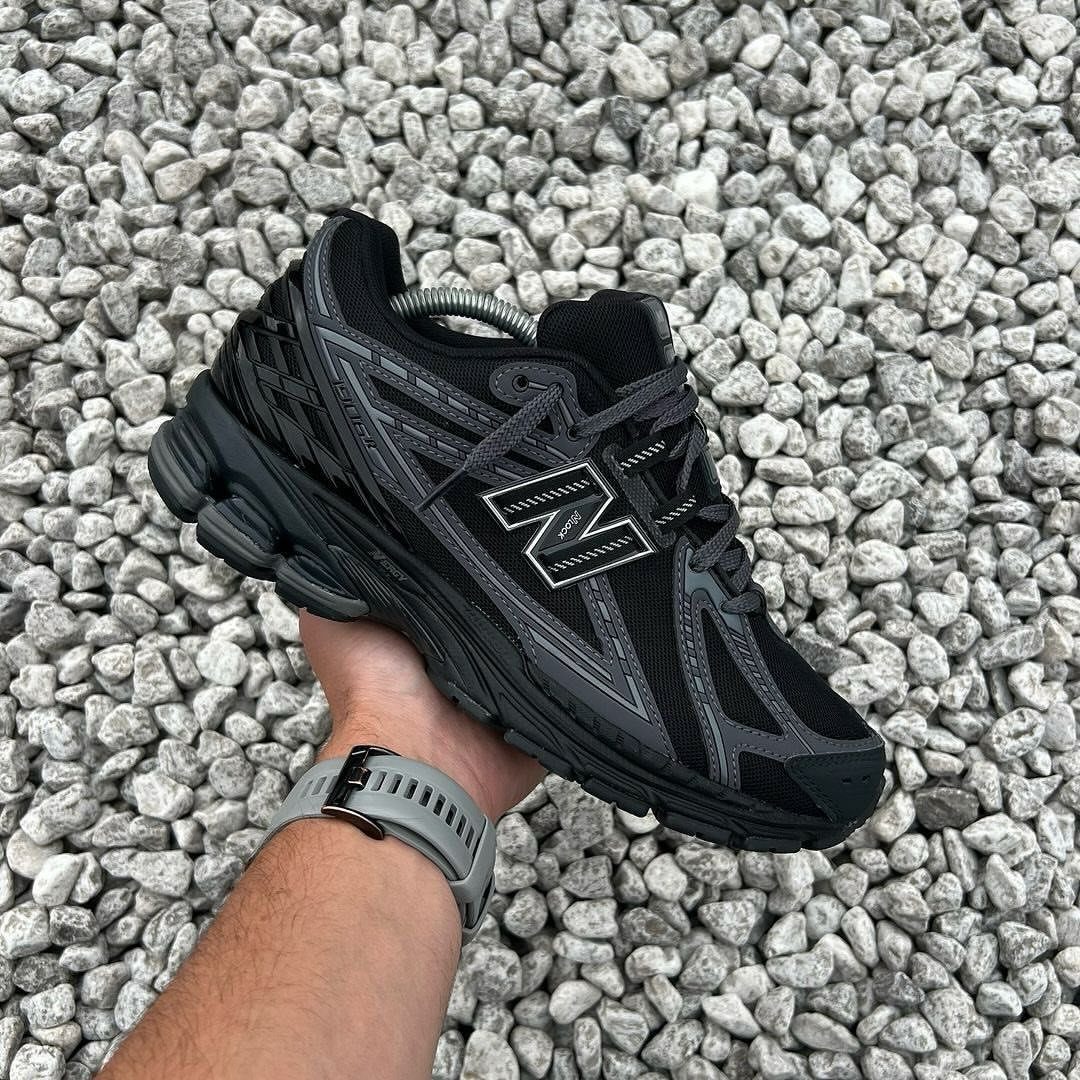Black New Balance 9060 sneaker held by a hand over grey gravel stones.