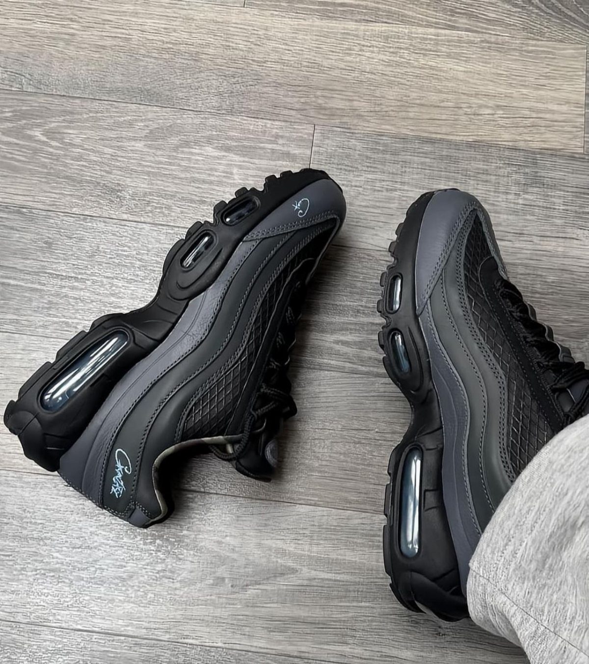 Black Nike Air Max Plus sneakers on a grey wood-grain floor.