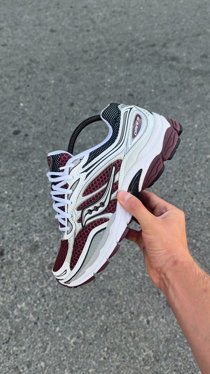 Saucony running shoe in white and burgundy held against a grey asphalt background