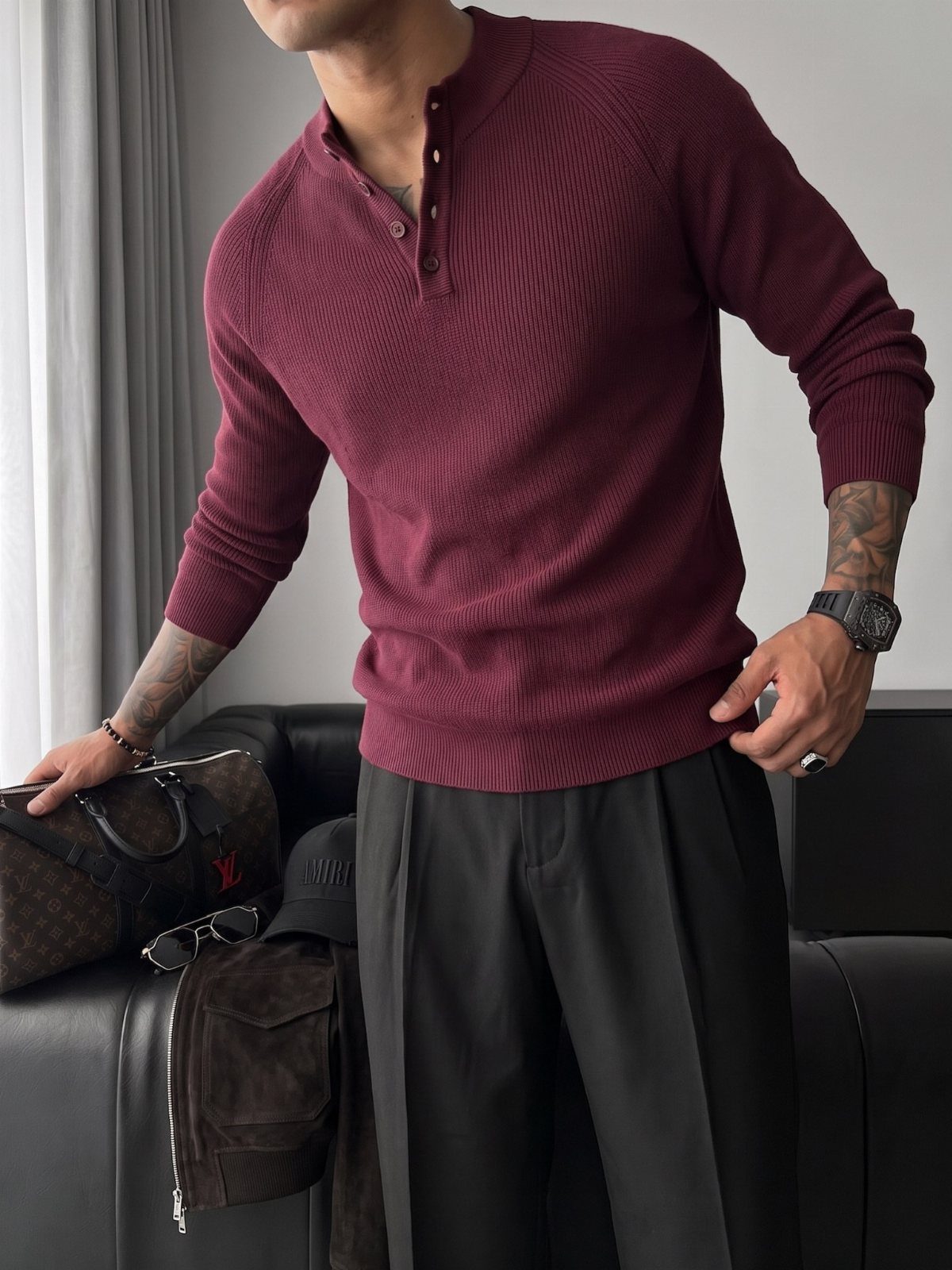 Man wearing a maroon ribbed henley shirt and black trousers standing by a sofa