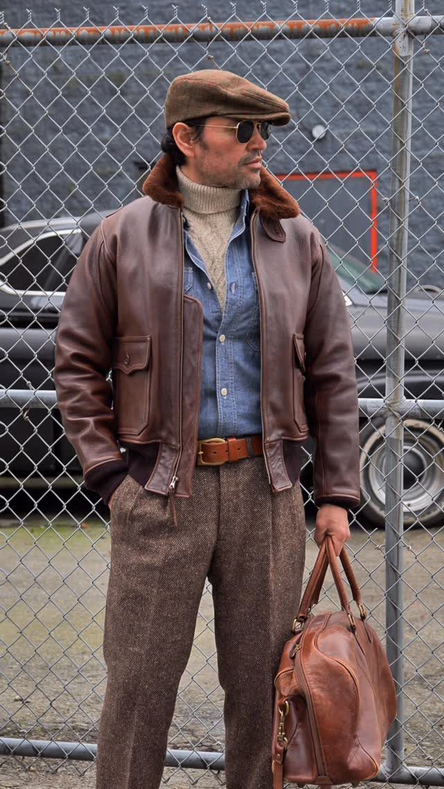 Man in leather jacket, denim shirt, and turtleneck posing by a chain-link fence.