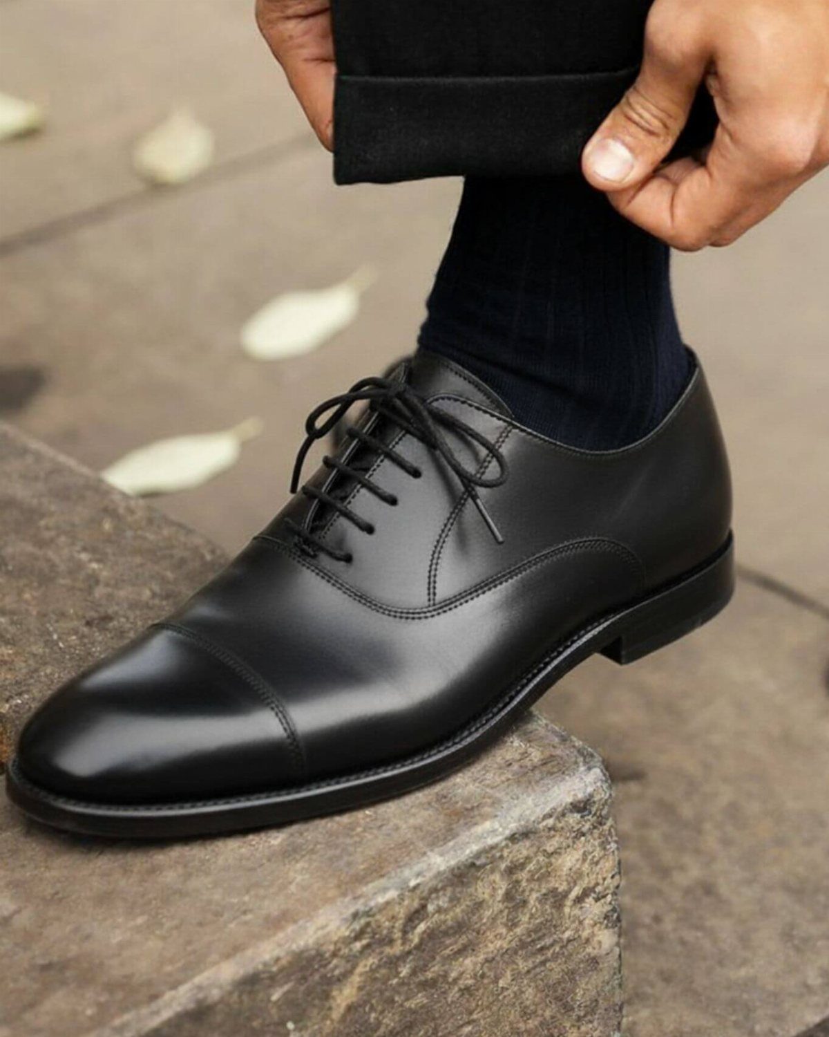 Black leather cap-toe oxford shoe on a stone step with dark trousers.