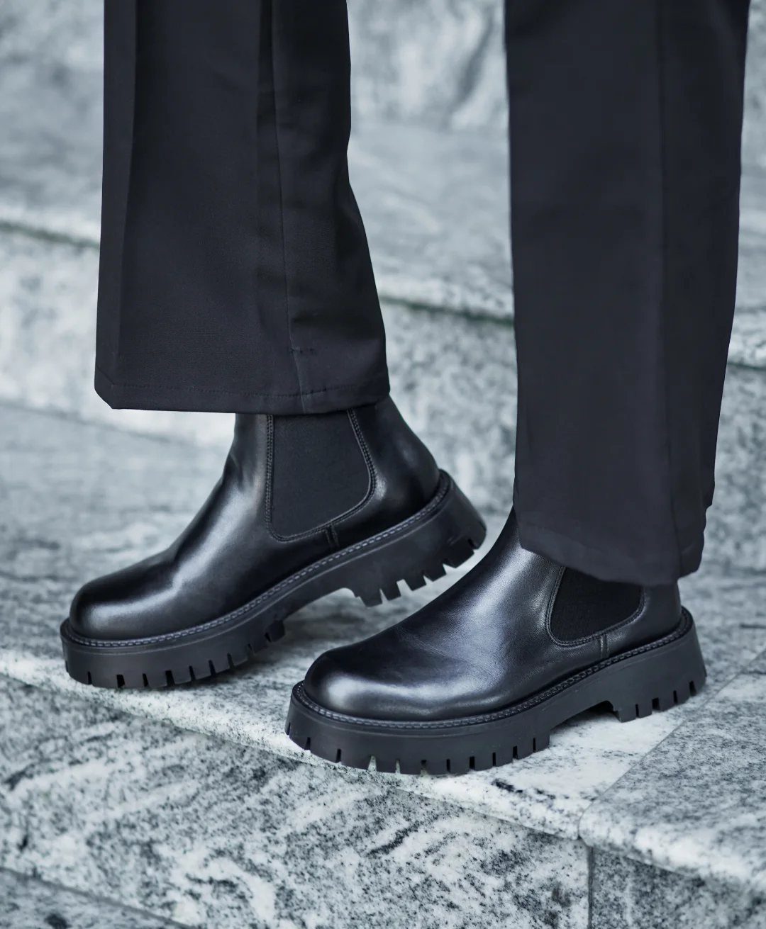 Black leather Chelsea boots with chunky soles worn with black trousers on marble.