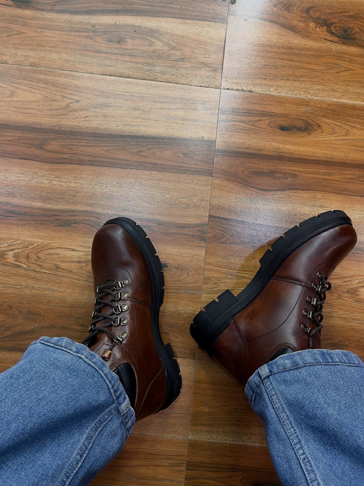 Brown leather lace-up boots worn with blue jeans on a wooden floor.