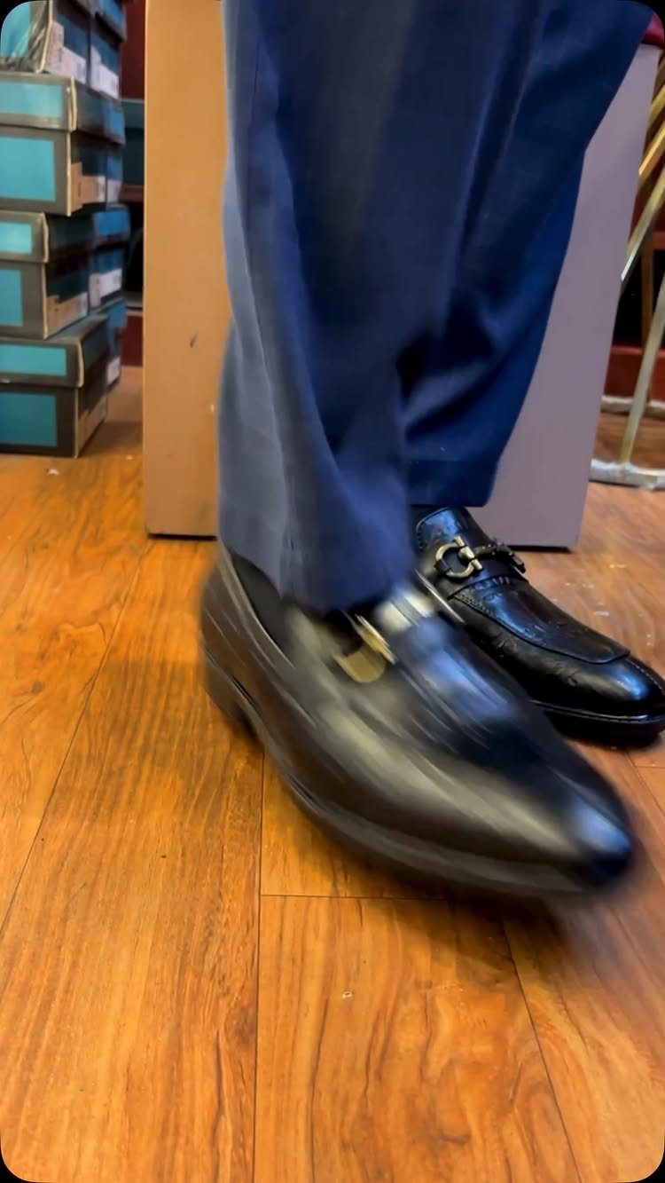 Black leather horsebit loafers worn with blue trousers on a wooden floor.
