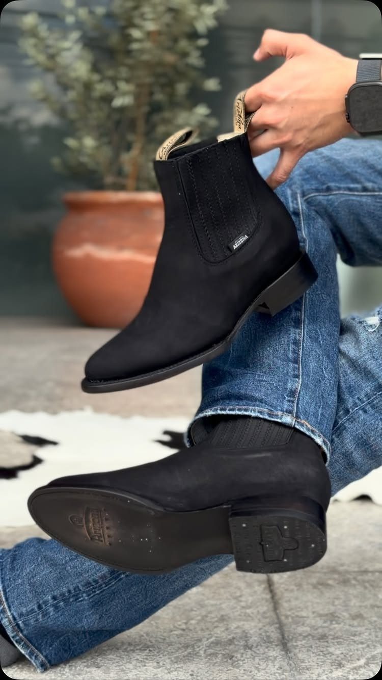 Black suede Chelsea boots paired with blue denim jeans on a person.