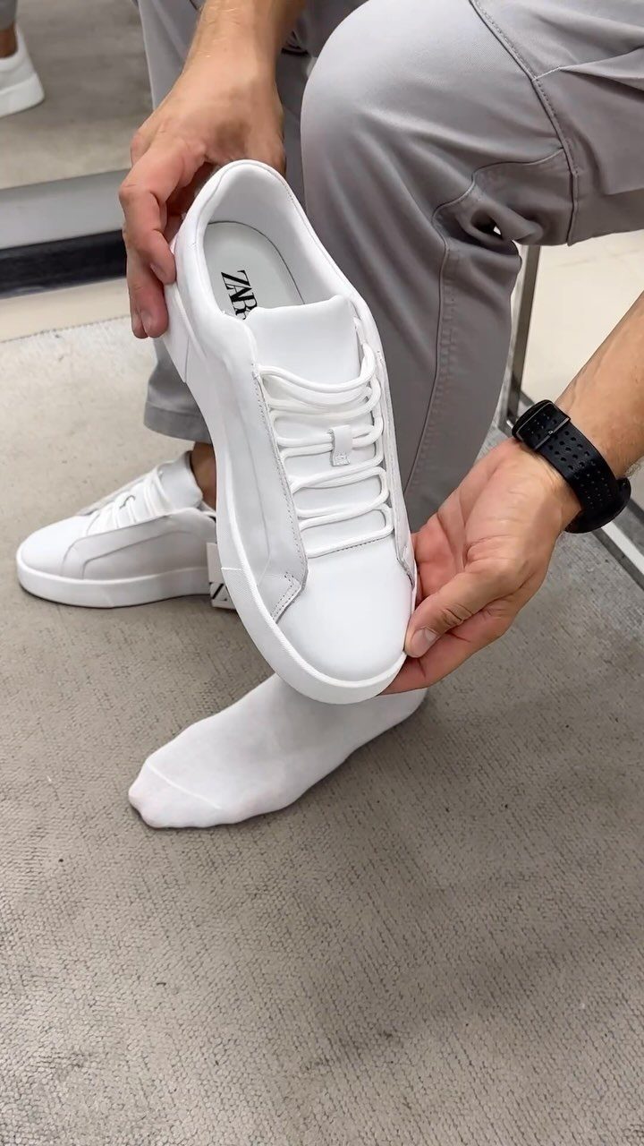 White leather sneakers held by a man wearing grey trousers and white socks.