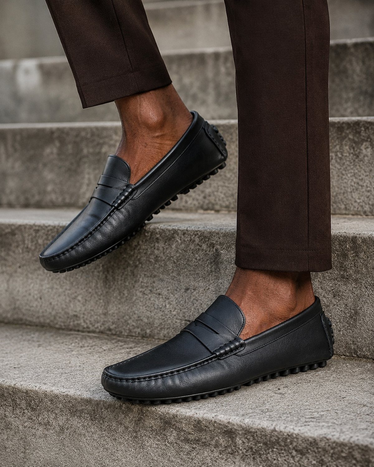 Black leather loafers worn with dark brown trousers on concrete stairs.