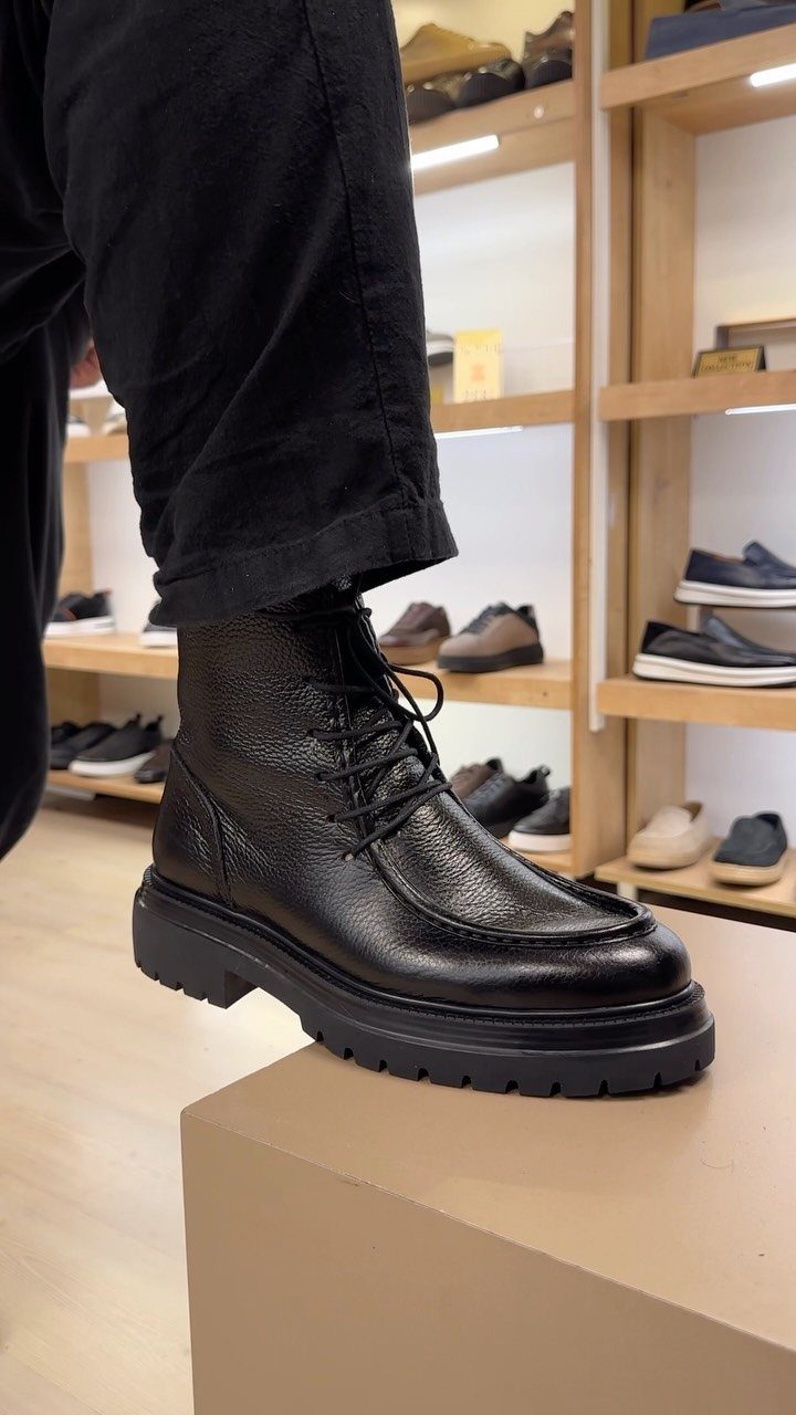 Black leather lace-up boots with thick soles worn with black trousers in a store.