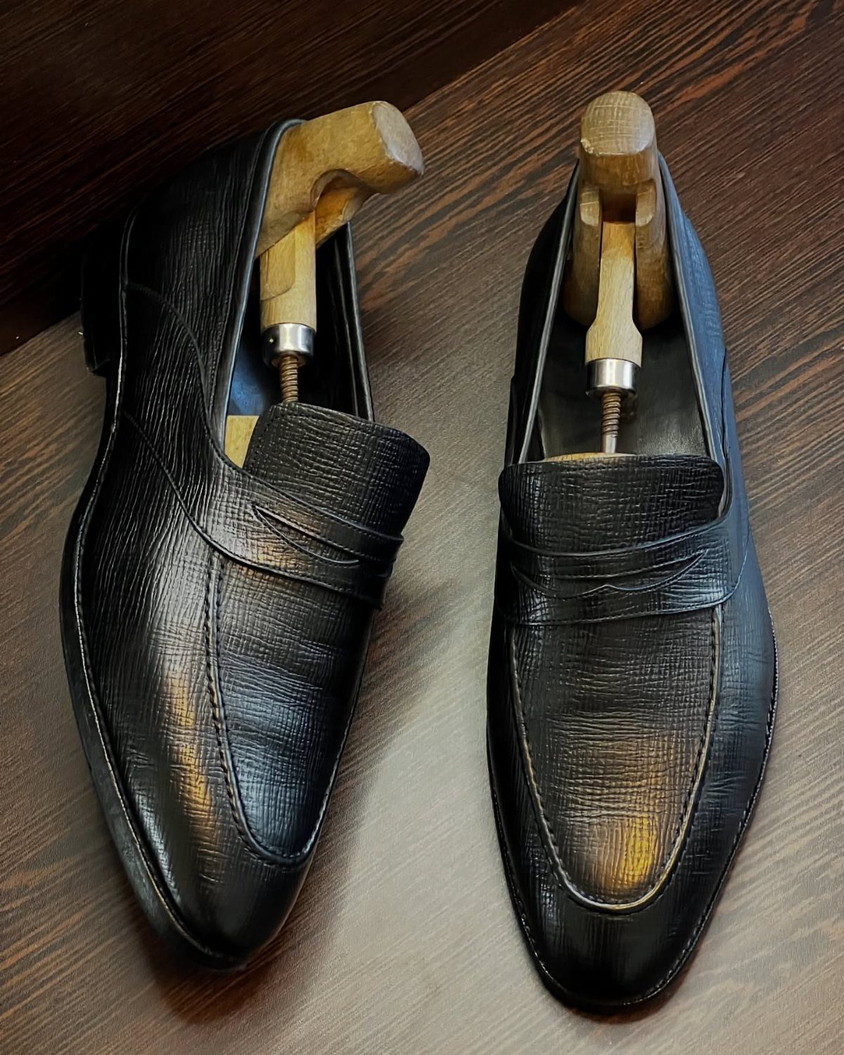 Black textured leather penny loafers with wooden shoe trees on a dark wood surface