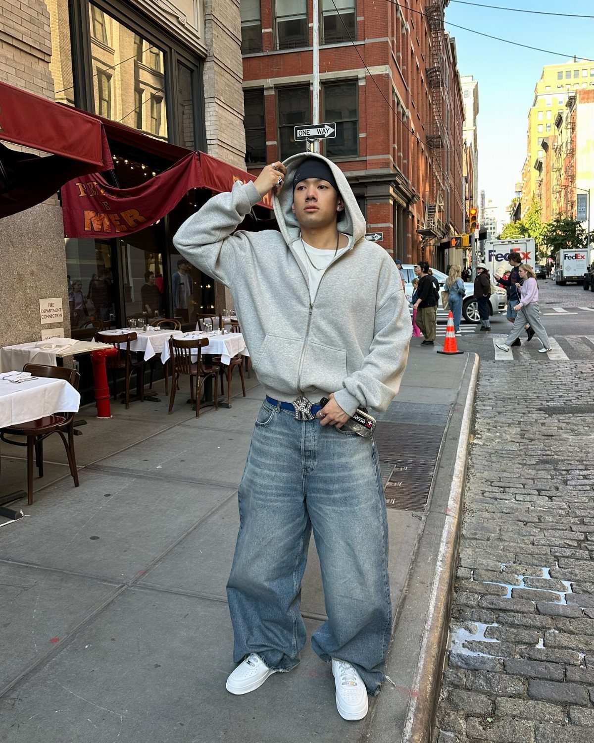 Man in grey hoodie and wide blue jeans wearing white athletic sneakers outdoors.
