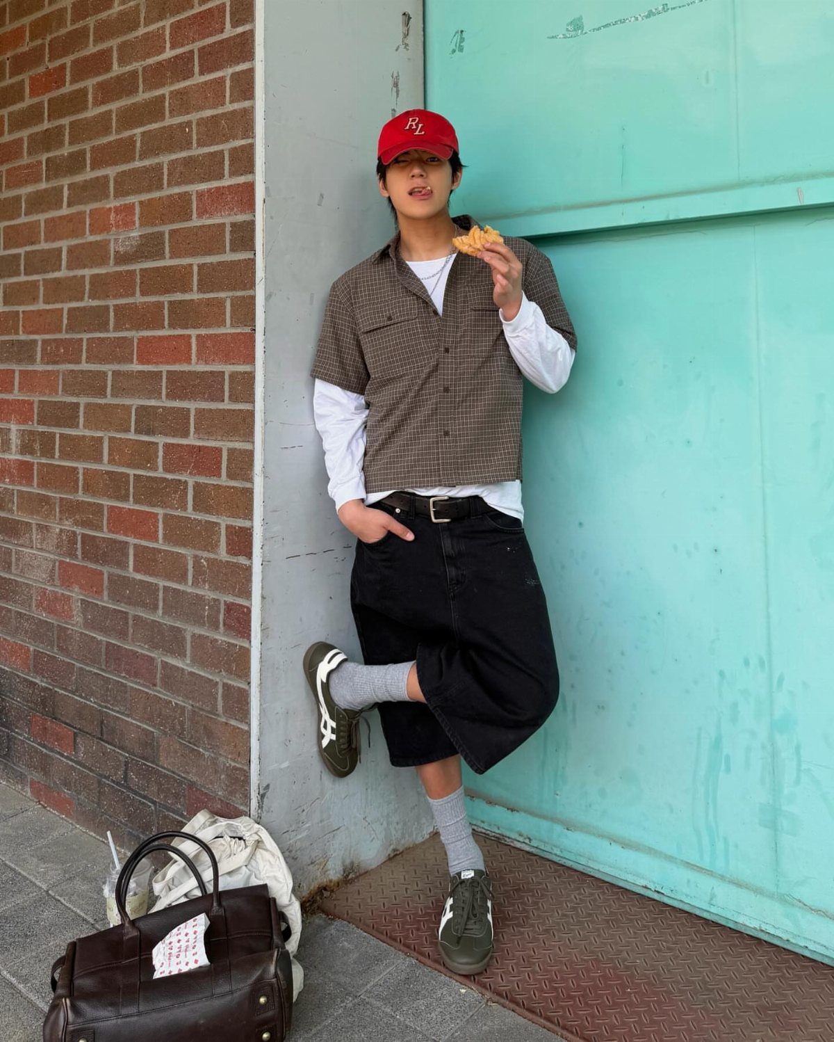 Person in baggy black shorts and grey Onitsuka Tiger sneakers leaning against a wall.