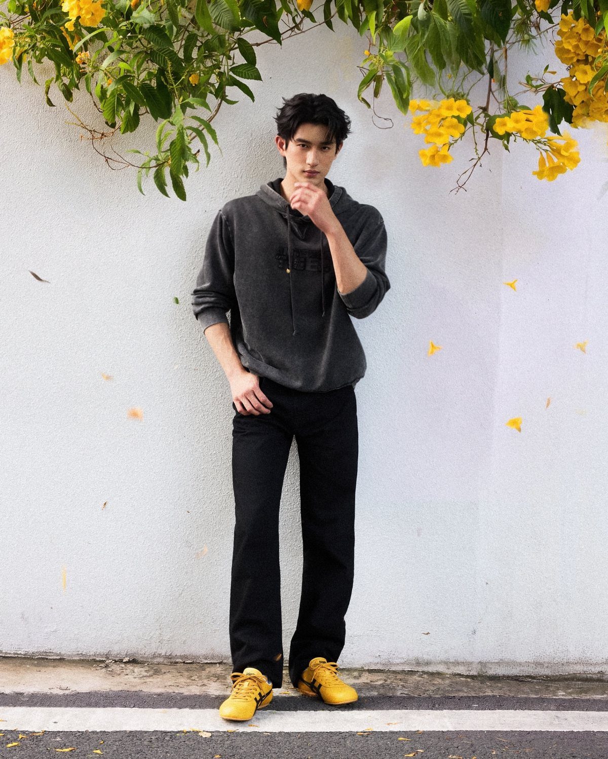 Man in grey hoodie and black trousers wearing bright yellow sneakers by a wall