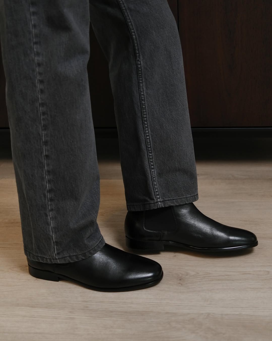Black leather Chelsea boots paired with dark grey denim jeans on wooden flooring.