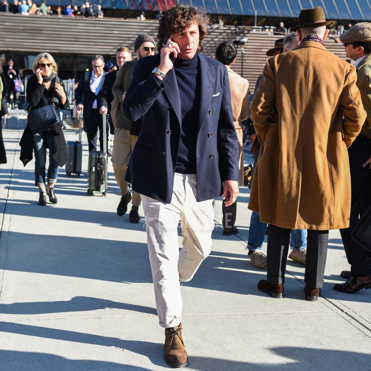Man wearing navy blazer, white trousers, and brown suede chukka boots walking outdoors.