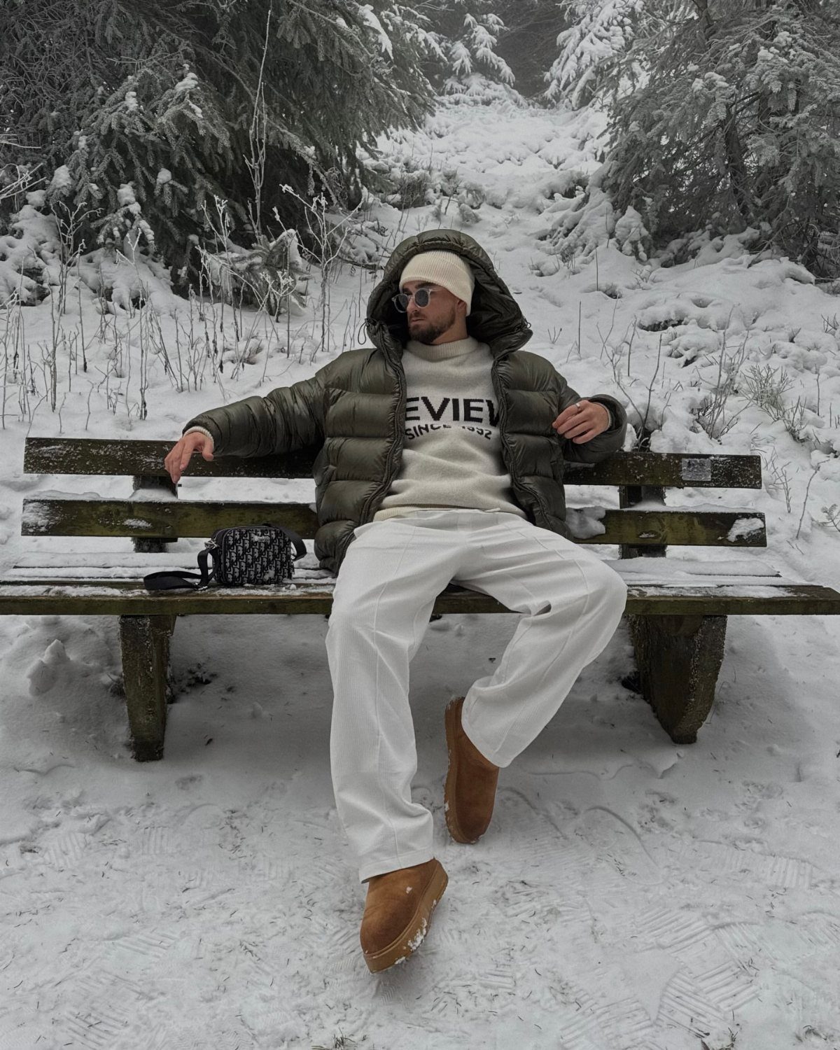 Man in a green puffer jacket and brown winter boots sitting on a snowy bench.
