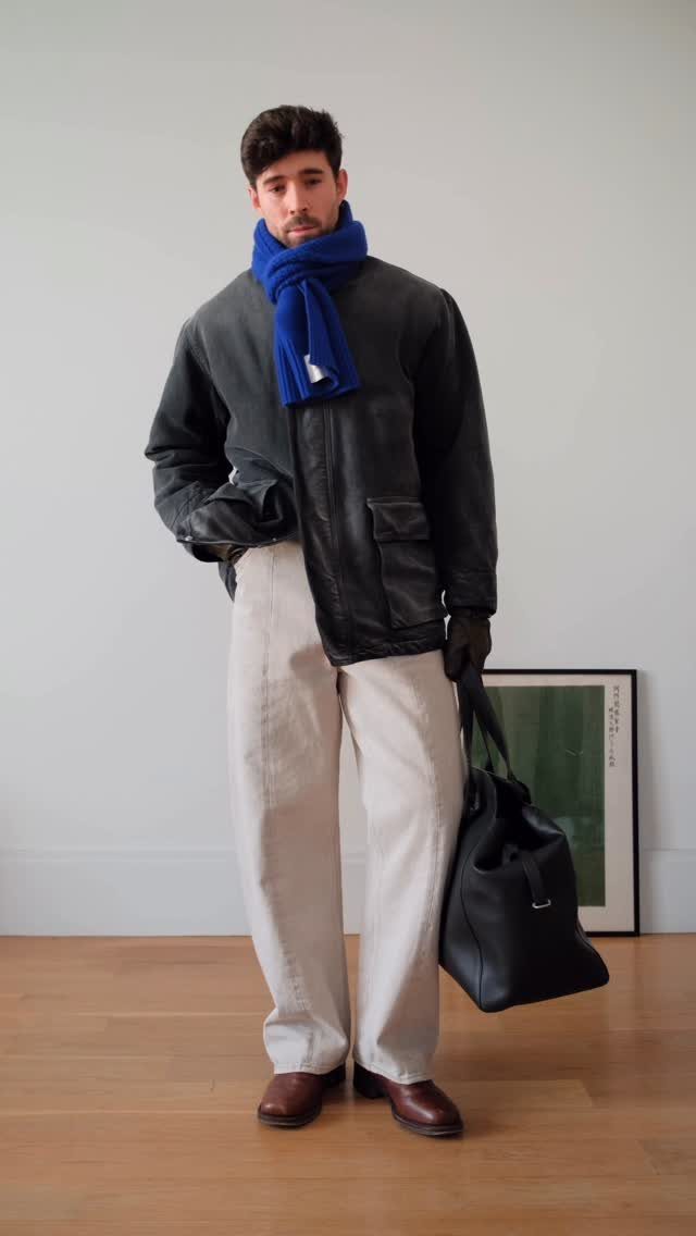 Man wearing a leather jacket, blue scarf, cream trousers, and brown winter boots.