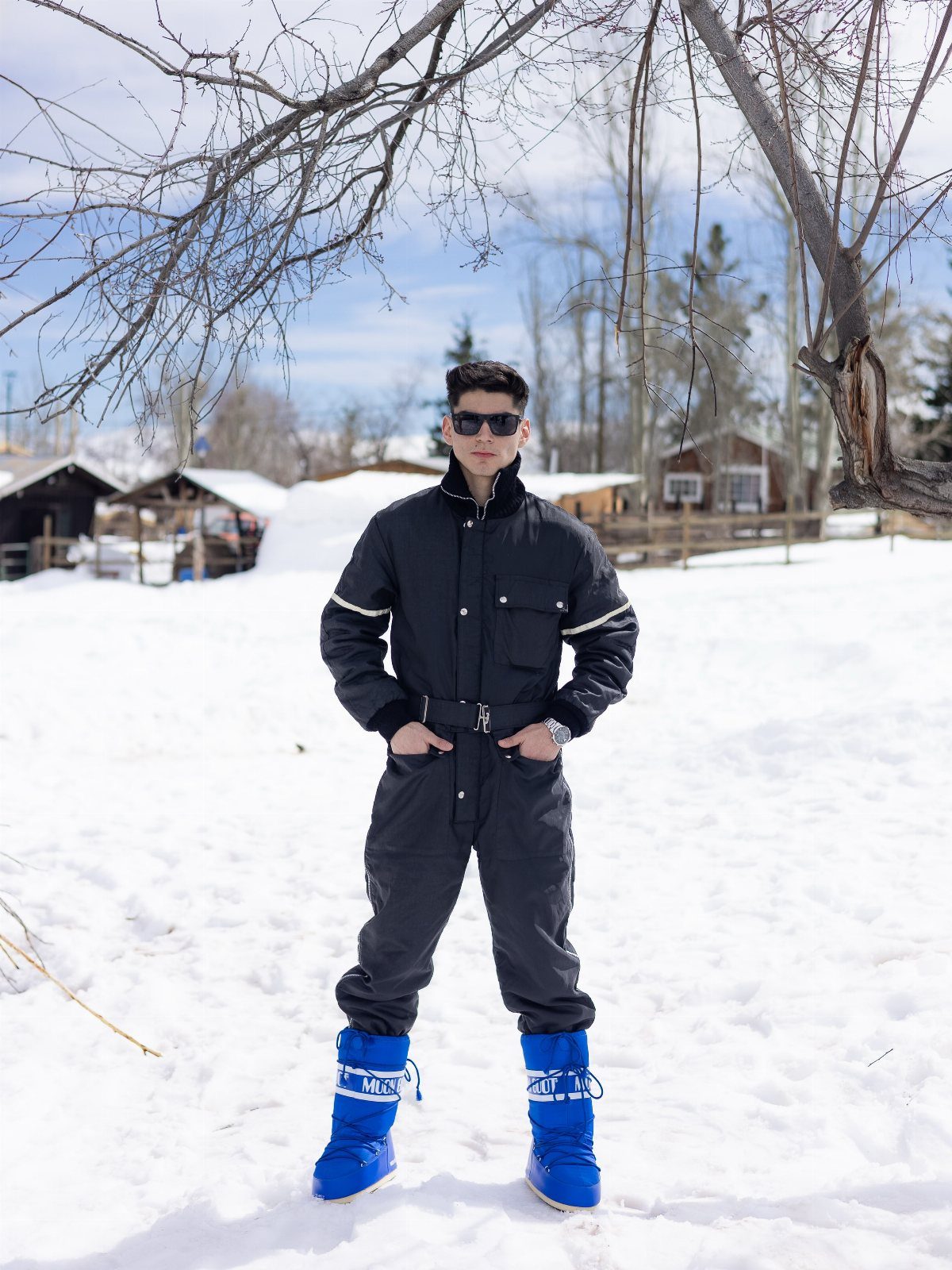 Man in black winter jacket and grey snow pants wearing bright blue snow boots.